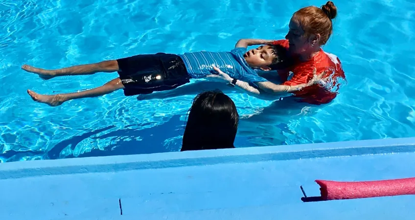 YMCA instructors support a student while they learn to float on their back during a swim lesson as part of the inaugural Learn to Swim program with GEHA in the summer 2022.