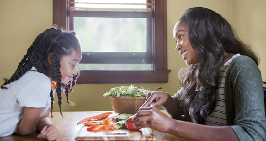 Mother and daughter smile while sitting at a table cutting up ingredients to put in a salad.