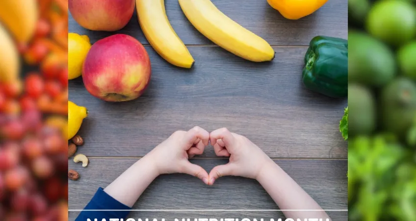 Child holding their hands in the shape of a heart on a table covered in a rainbow of fruit and vegetables. Text: "National Nutrition Month".