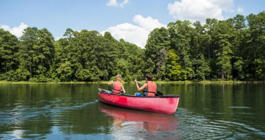 Two kids in lifejackets paddle a red canoe across a lake toward a tree-covered shoreline.