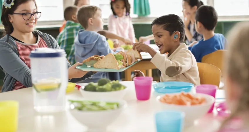 A group of children eat around a table. The table is filled with vegetables and other health foods.