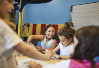 Girls sit at a table smiling as they color with markers, while other kids are playing in the background.