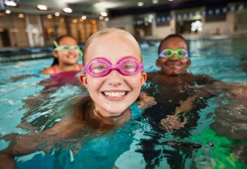 Three kids in the water wearing goggles and smiling at the camera.