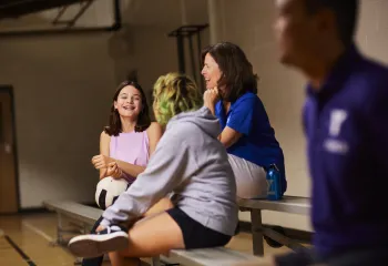 Two volleyball players sit on the bleachers and talk with a YMCA associate.