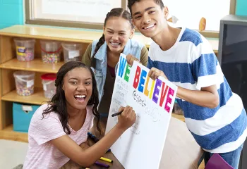 Three teenagers smile at the camera while holding a poster that says "We Believe"