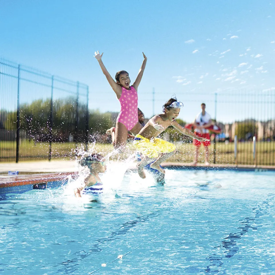 3 children smiling as they jump into outdoor pool at Greater Kansas City YMCA with a lifeguard watching them in the distance.