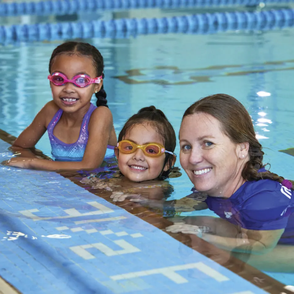 2 swim students smiling with Y instructor while hanging on the pool edge.