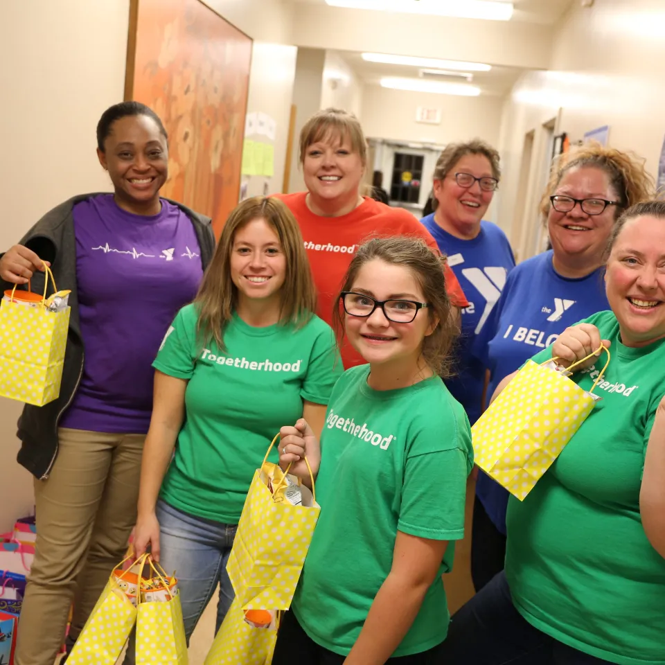 Group of adults and teenagers hold bags they're giving out while wearing Togetherhood t-shirts at YMCA Volunteer event.