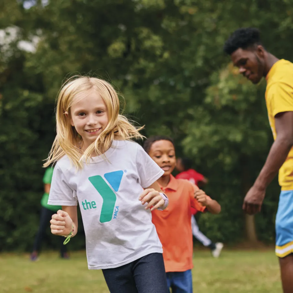 Smiling children wearing Greater Kansas City YMCA shirts running and playing in field during a summer day camp.