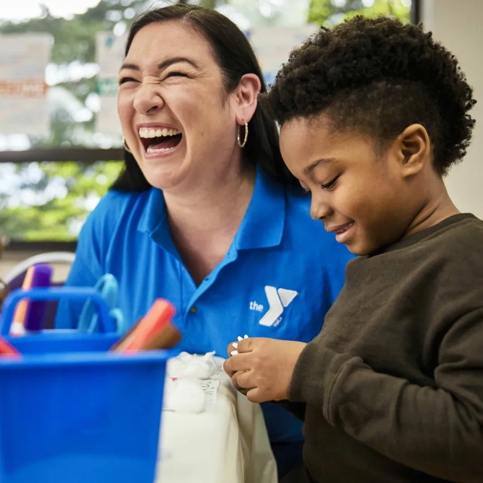 A Y staff member smiles and laughs while helping a young child on an art project involving markers 