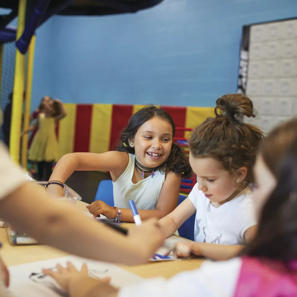 Girls sit at a table smiling as they color with markers, while other kids are playing in the background.