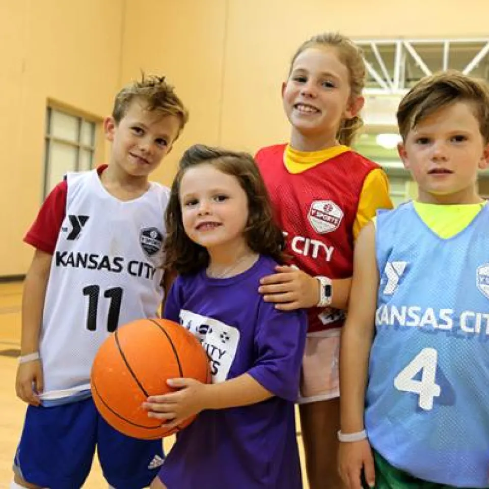 A group of children stand smiling holding a basketball at the North Kansas City YMCA indoor basketball court.