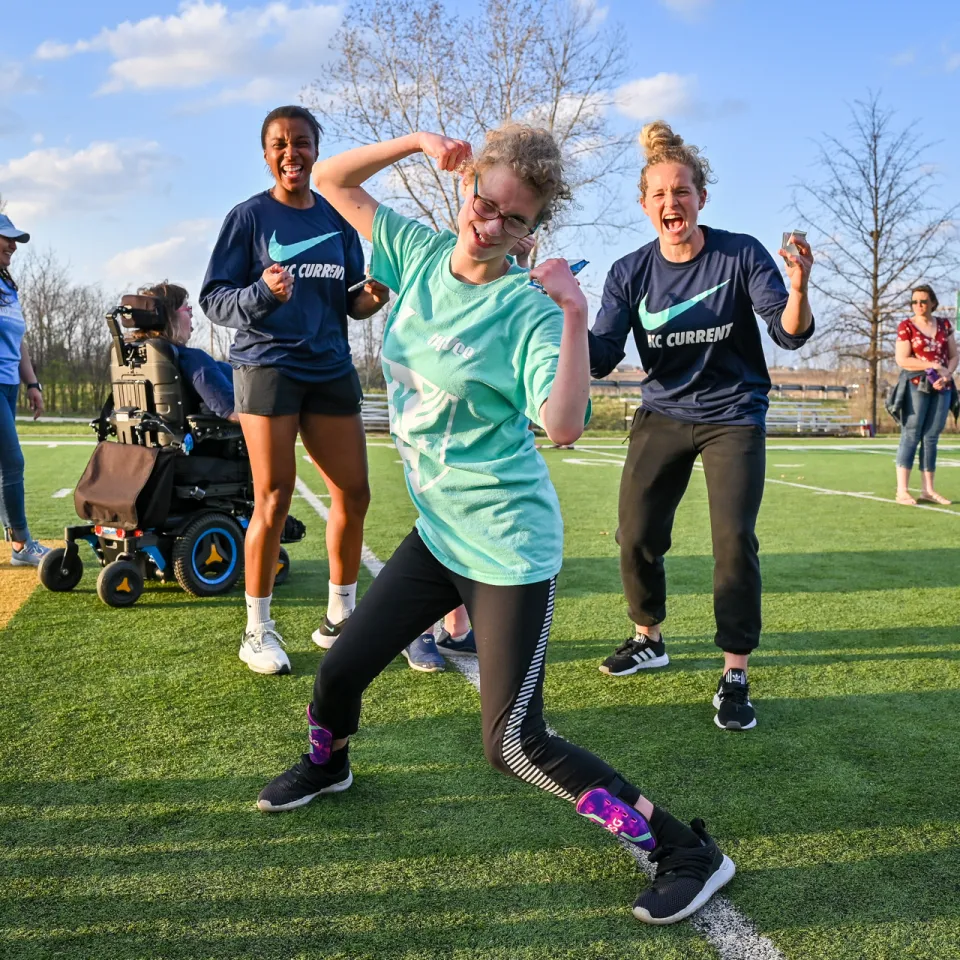 KC Current YMCA Challenger Soccer player flexes for the camera, two KC Current Soccer players cheer behind her.