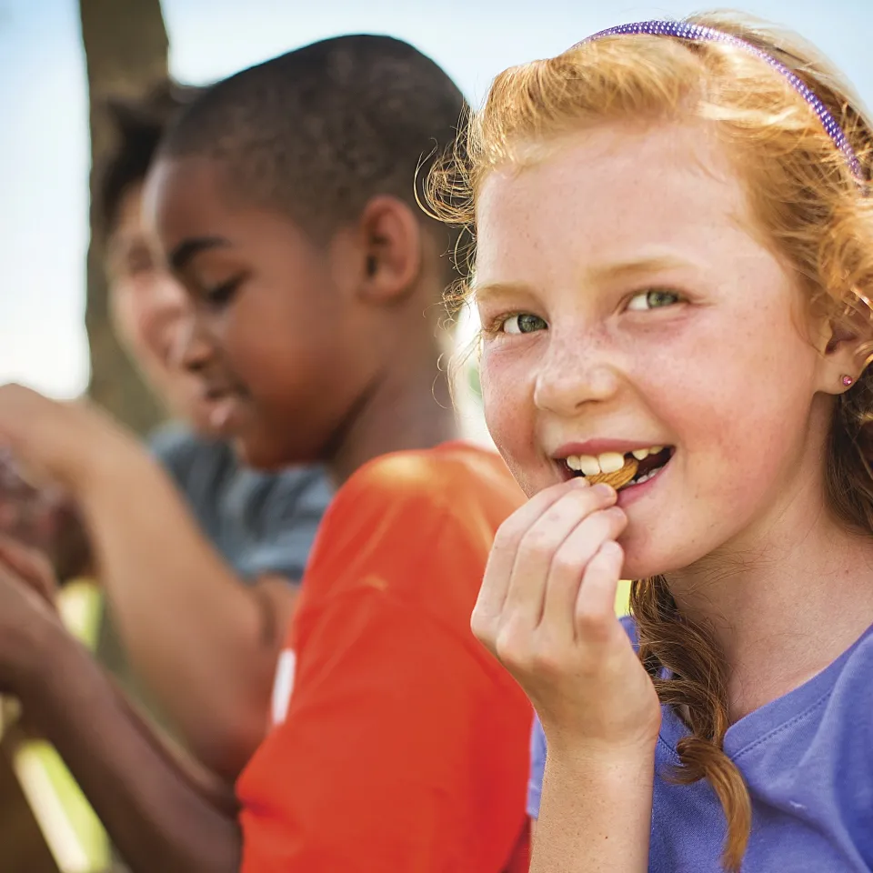 Photo of girl eating snack, smiling, with two boys in the background