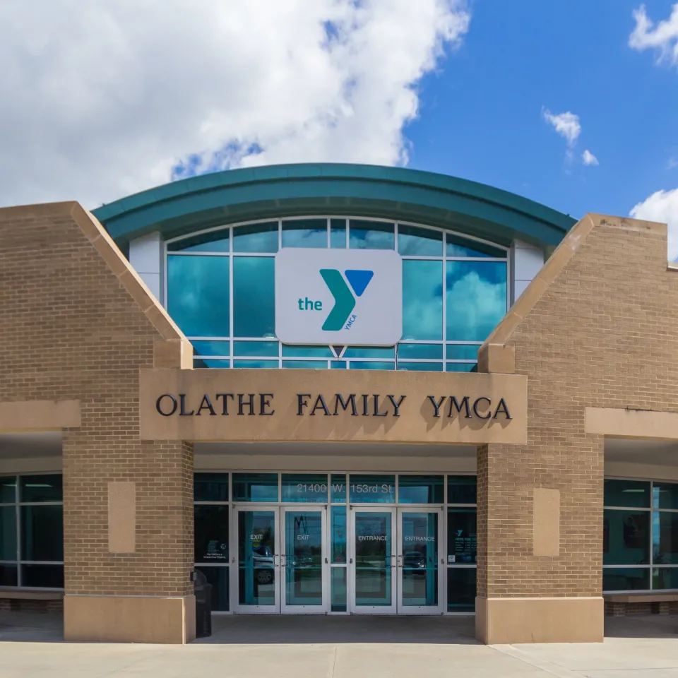 Exterior of the Olathe Family YMCA. Buildings front entrance looks like a brick colosseum with "Olathe Family YMCA" in the very center. Beyond the brick is floor to ceiling glass with the Y logo displayed in the center of the glass. 