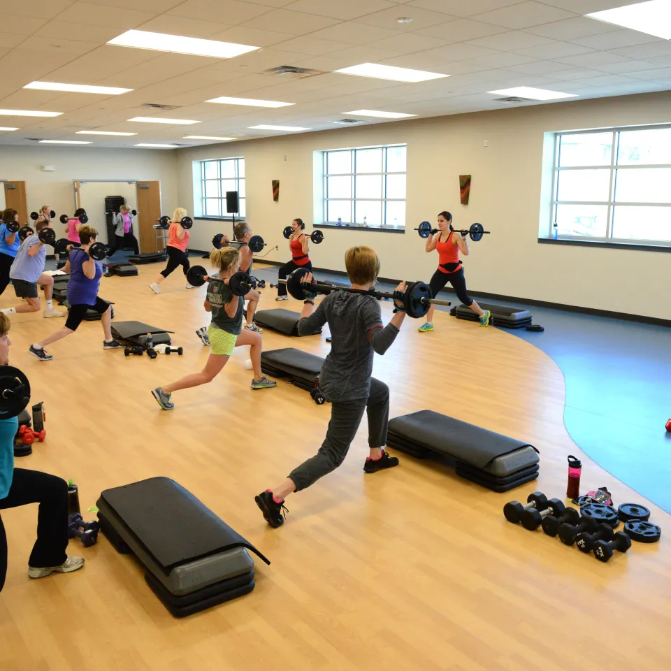 Group exercise class participants are led by a Y instructor with weights. They are challenging their strength and balance with weighted forward lunges.