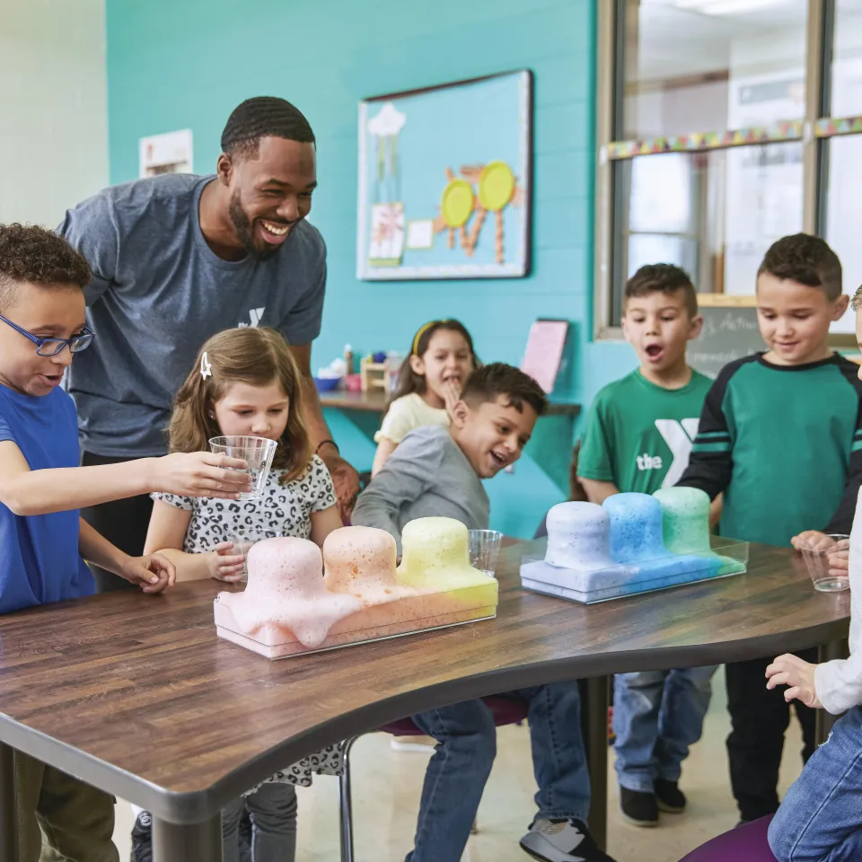 Y Staff Member oversees a table full of students in a science experiment where a rainbow of foam is overflowing from beakers, during a No School Day.