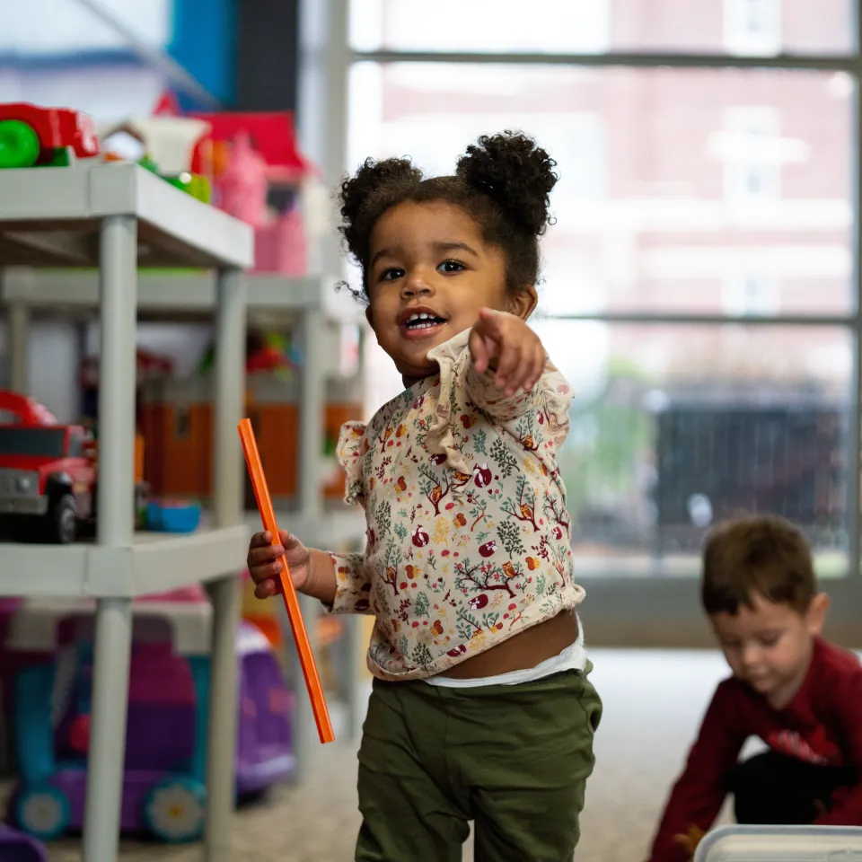 Child pointing toward the camera with another child playing behind her in a Kids Zone at a Y center.