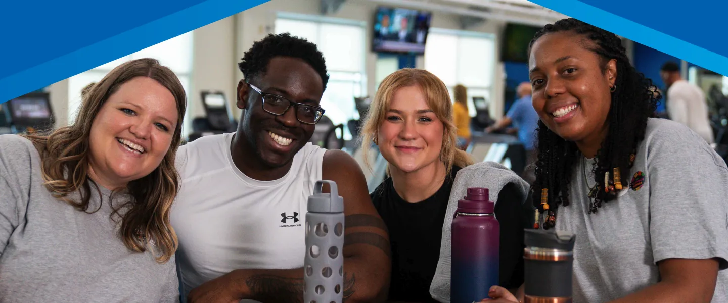 Four young adults with water bottles socialize in the lobby of the Y, smiling, after a workout