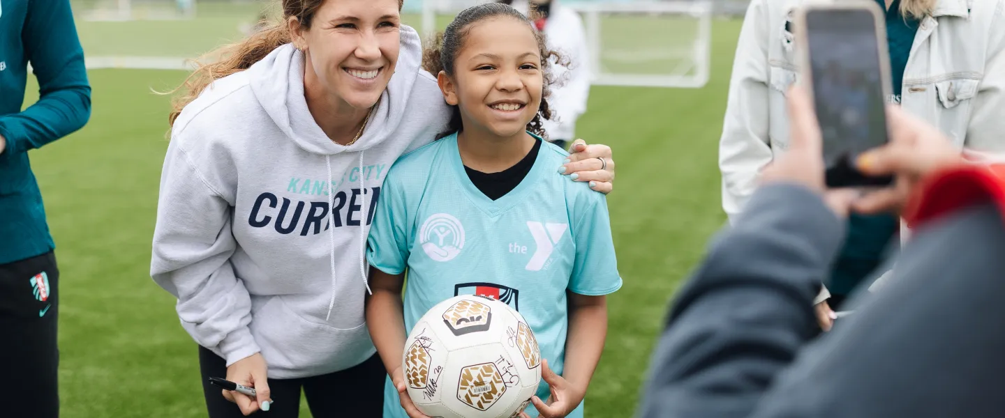 KC Current soccer player poses with girl wearing YMCA KC Current soccer jersey and holding autographed soccer ball