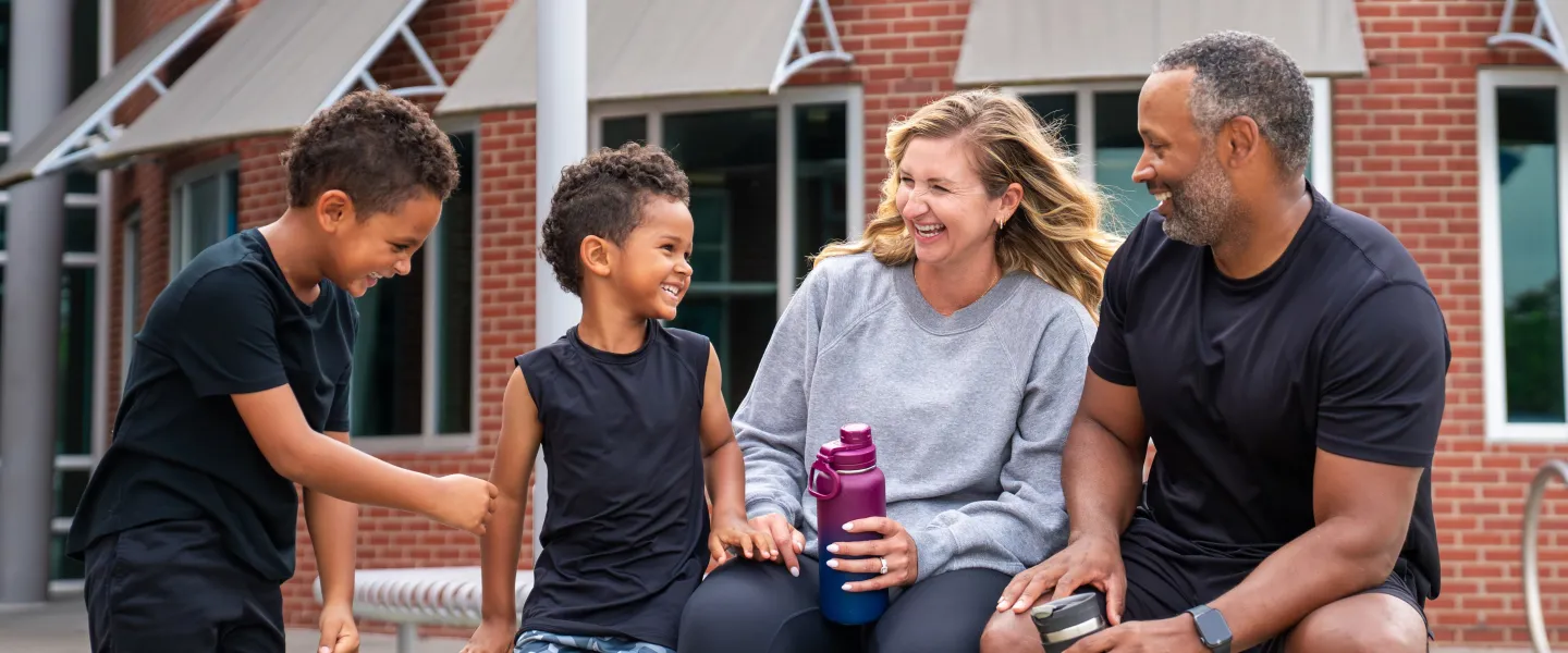 Photo of two boys, mom and dad sitting on bench outside the YMCA holding water bottles and smiling