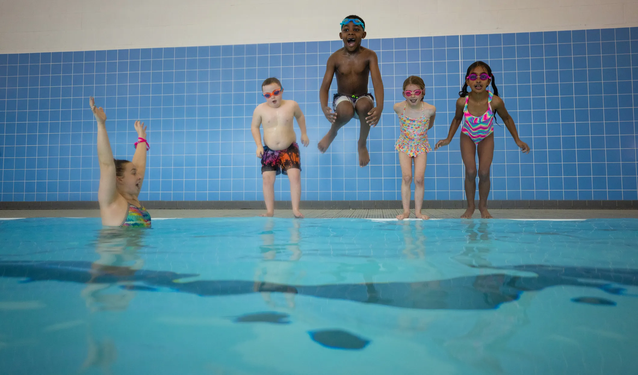 Children getting ready to jump into an indoor pool with their instructor in the pool cheering them on.
