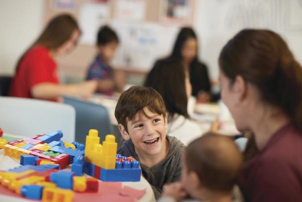 A young boy sits at a table with large Legos on it. He is smiling at an adult woman, who has her back turned to the camera