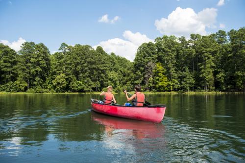 Two kids in lifejackets paddle a red canoe across a lake toward a tree-covered shoreline