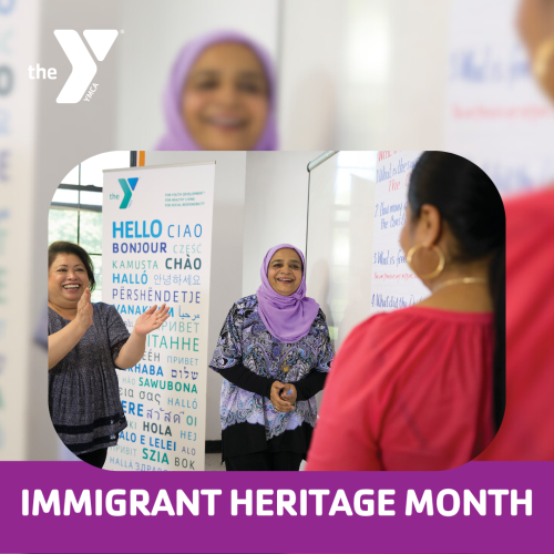 Two people smile and clap their hands to a person whose back is facing the camera. They are in front of a YMCA sign saying "hello" in many different languages. Text, "Immigrant Heritage Month"