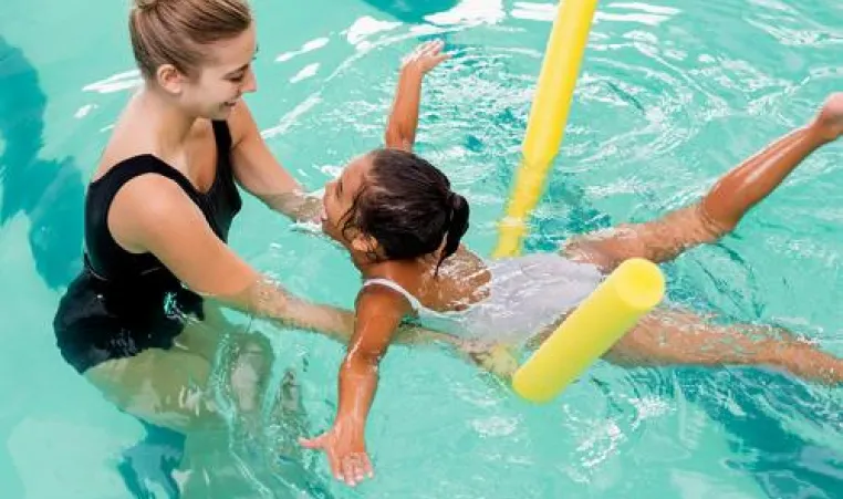 Young girl floating on a pool noodle while swimming toward a swim instructor. 