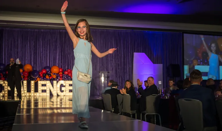 Challenger athlete Maddie Richardson struts down the runway in a floor length blue gown and a cute white purse. The audience is smiling and clapping.