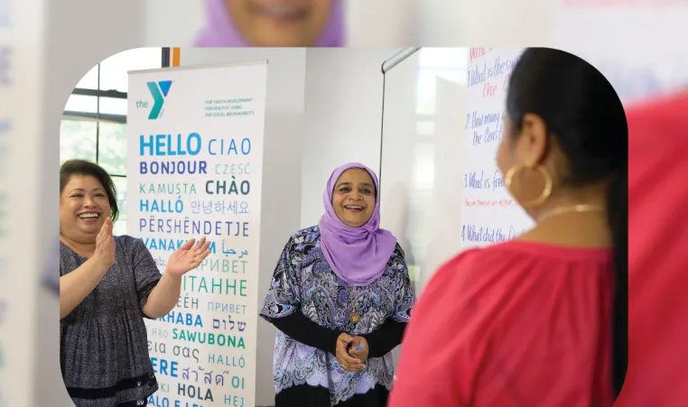 Kansas City YMCA graphic featuring two women smiling and clapping their hands to a person whose back is facing the camera. They are in front of a YMCA sign saying "hello" in many different languages. There's a bottom text banner saying "Immigrant Heritage Month".