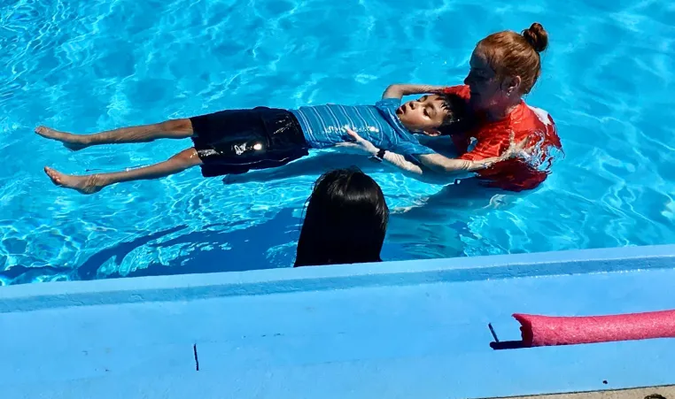 YMCA instructors support a student while they learn to float on their back during a swim lesson as part of the inaugural Learn to Swim program with GEHA in the summer 2022.