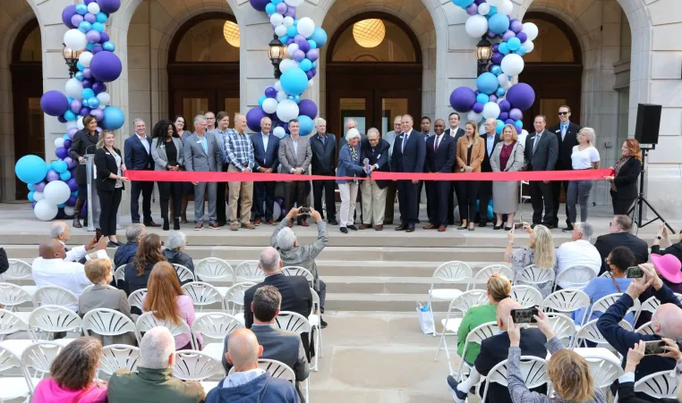 Kirk family uses large scissors to cut red ribbon at ribbon cutting ceremony. Family is surrounded by representatives from donors, Y leadership and community leaders. Members of the audience are seated, many taking photos on their cell phones.. 