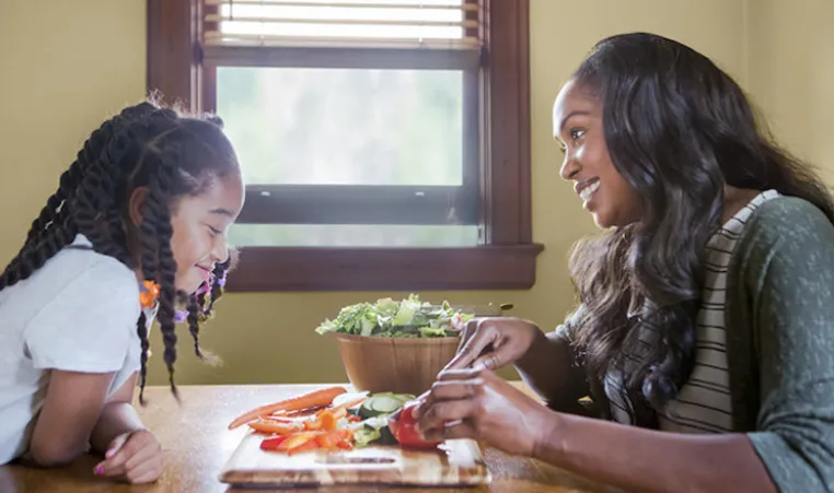 Mother and daughter smile while sitting at a table cutting up ingredients to put in a salad.