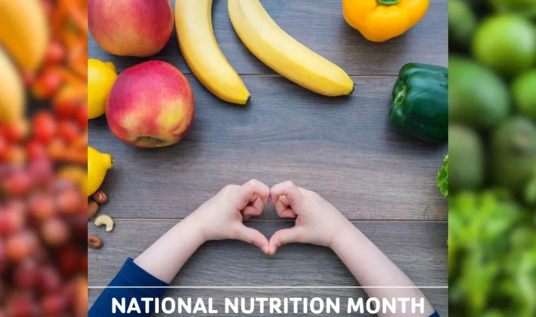 Child holding their hands in the shape of a heart on a table covered in a rainbow of fruit and vegetables. Text: "National Nutrition Month".