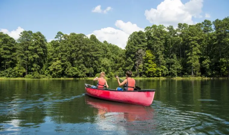 Two kids in lifejackets paddle a red canoe across a lake toward a tree-covered shoreline.