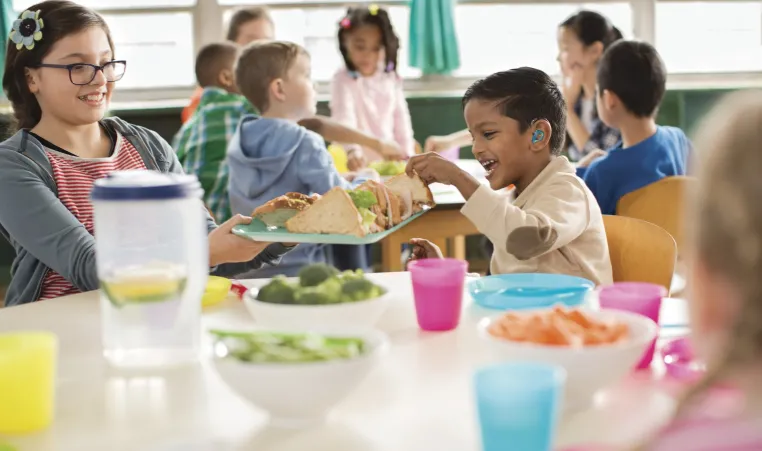 A group of children eat around a table. The table is filled with vegetables and other health foods.