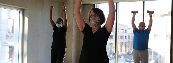 Group exercise class, three participants lift hand weights with arms extended straight up